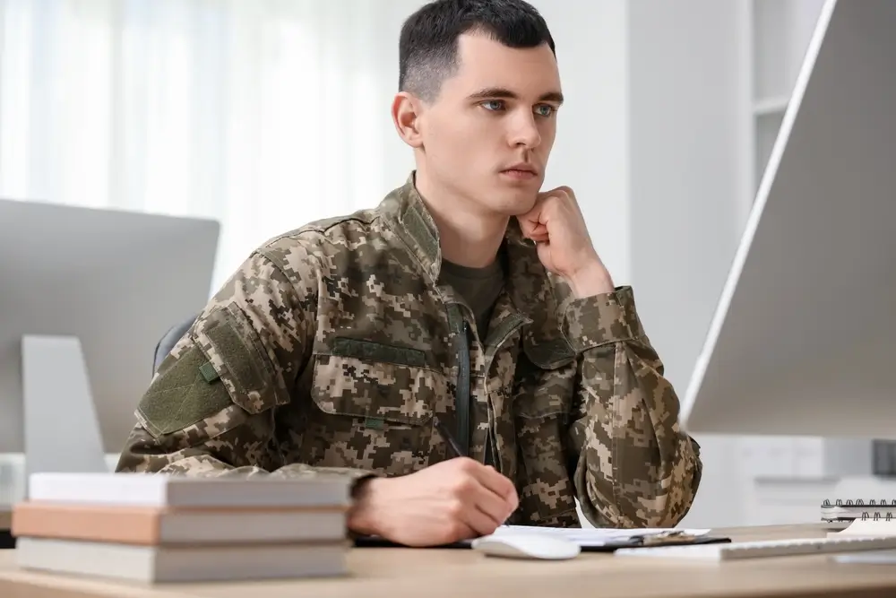 Estudiante con uniforme militar preparando sus oposiciones frente al ordenador con libros y material de estudio.