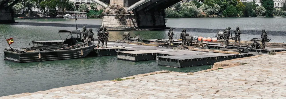 Infantes de marina españoles operando en una plataforma flotante junto a una embarcación en un río bajo un puente.