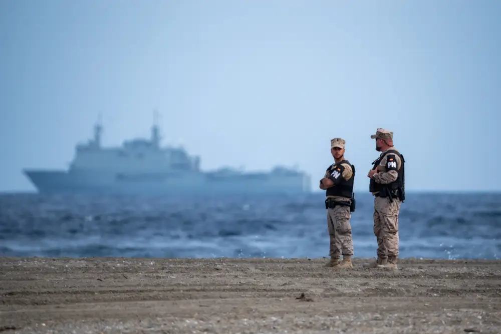 Militares de la Policía Naval con uniformes de camuflaje en la playa y un buque de la Armada al fondo.