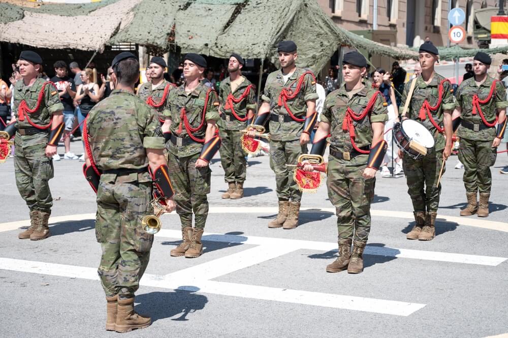 Soldados españoles en formación durante un desfile militar, vistiendo uniformes de camuflaje y cordones rojos de gala.