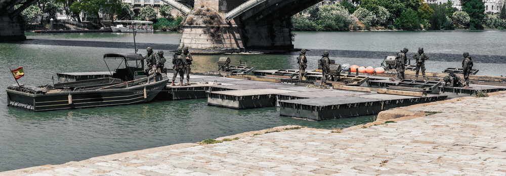 Infantes de marina españoles operando en una plataforma flotante junto a una embarcación en un río bajo un puente.
