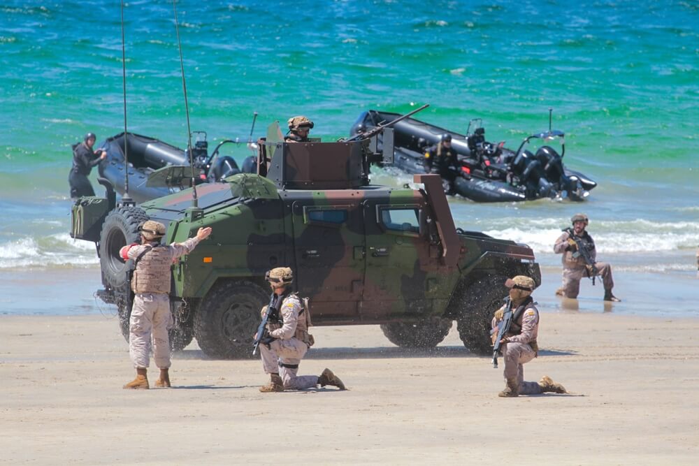Candidatos a militares realizando un ejercicio táctico de resistencia con vehículos blindados en una playa.