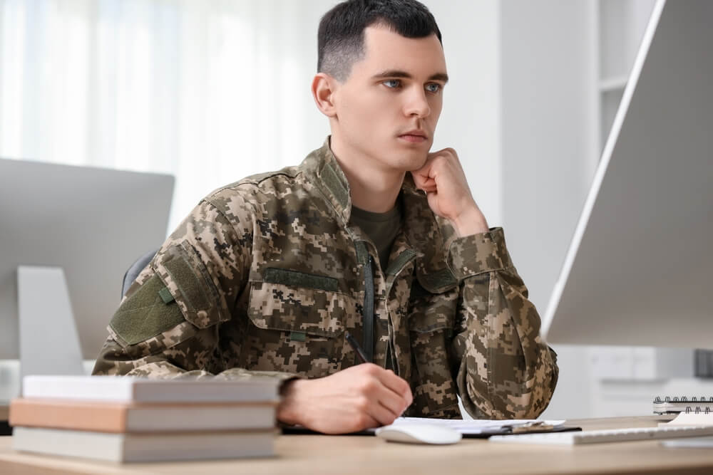 Estudiante con uniforme militar preparando sus oposiciones frente al ordenador con libros y material de estudio.