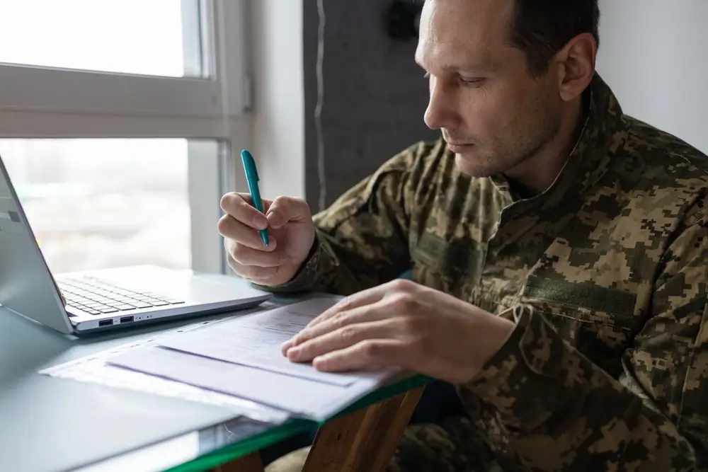 Militar firmando documentos y realizando trámites administrativos oficiales en una oficina con un ordenador portátil.