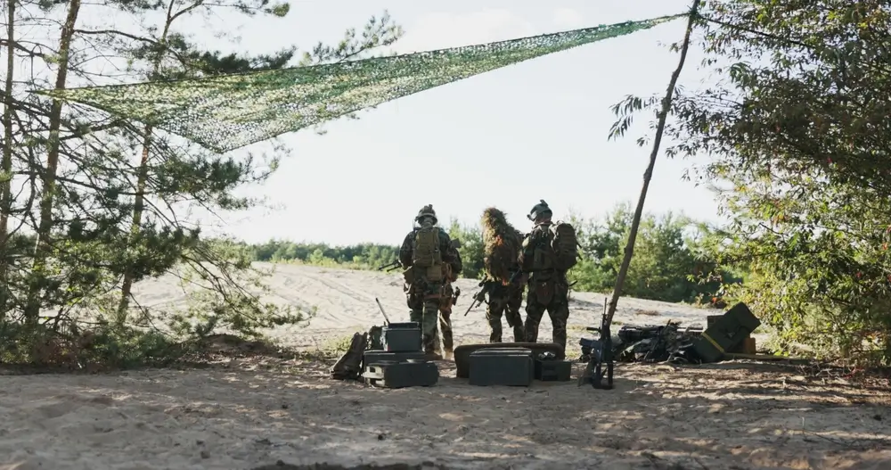 Soldados con equipo táctico bajo una red de camuflaje durante una instrucción en un campo de maniobras militar.