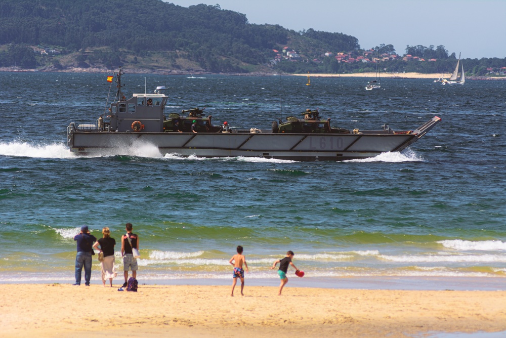 Lancha de desembarco L610 de la Armada Española navegando cerca de la costa frente a bañistas en la playa.