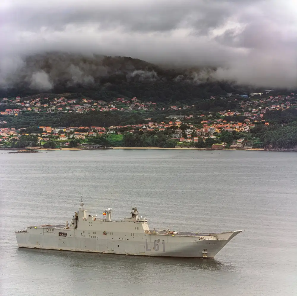 Buque L-61 Juan Carlos I de la Armada Española en una bahía frente a una localidad costera y montañas con niebla.