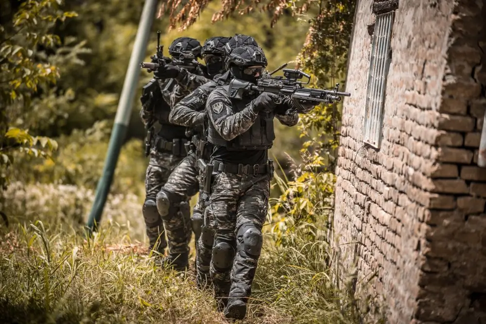 Soldados del MOE con equipo táctico de operaciones especiales avanzando en formación junto a un muro de ladrillo.