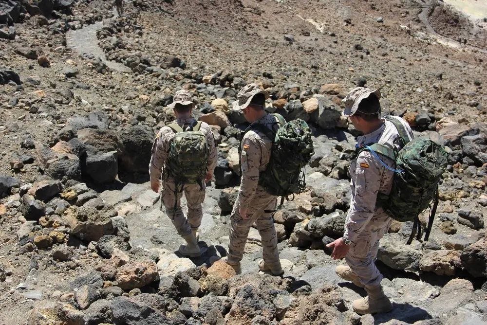Soldados en instrucción de montaña con equipo completo y mochilas tácticas avanzando por un terreno rocoso y escarpado.