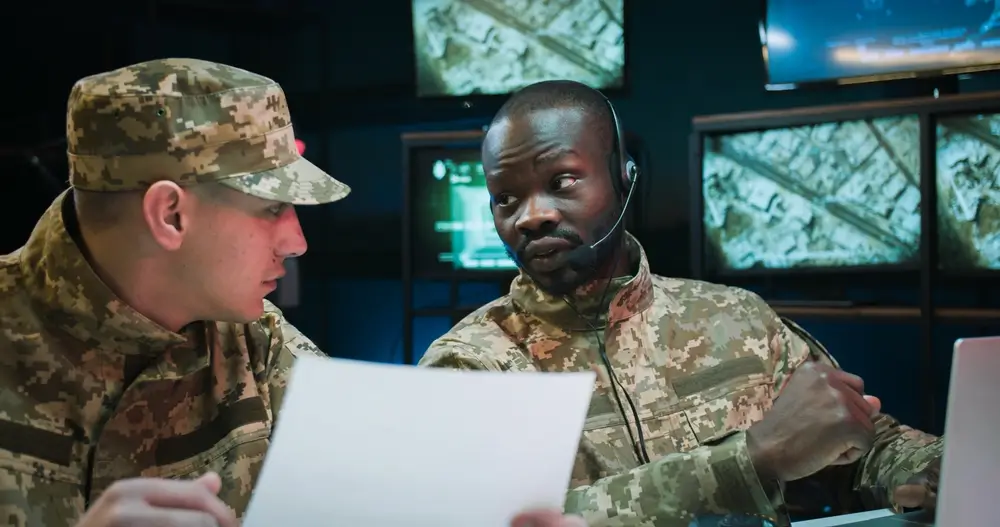 Dos oficiales del ejército analizando un expediente en un despacho militar con pantallas de control al fondo.