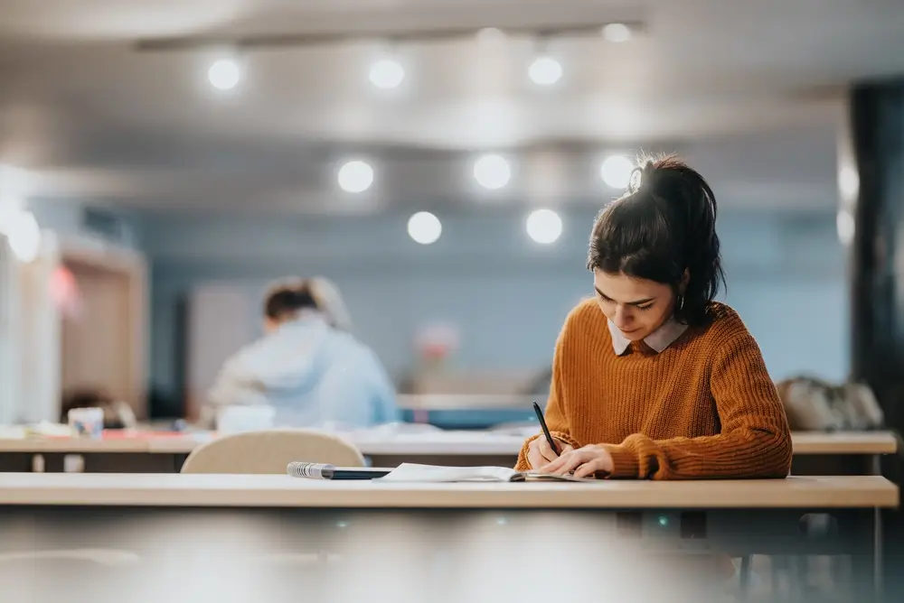 Estudiante en su escritorio preparando oposiciones mientras toma notas concentrada en un cuaderno.